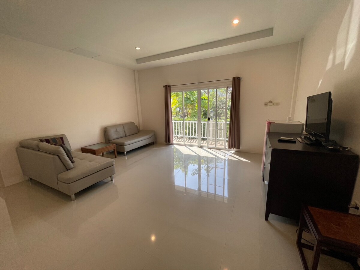 An airy living space features large glass doors opening to a balcony, allowing natural light to brighten the room. Two comfortable sofas are arranged with a wooden coffee table at the center. A television is positioned on a cabinet against the wall.