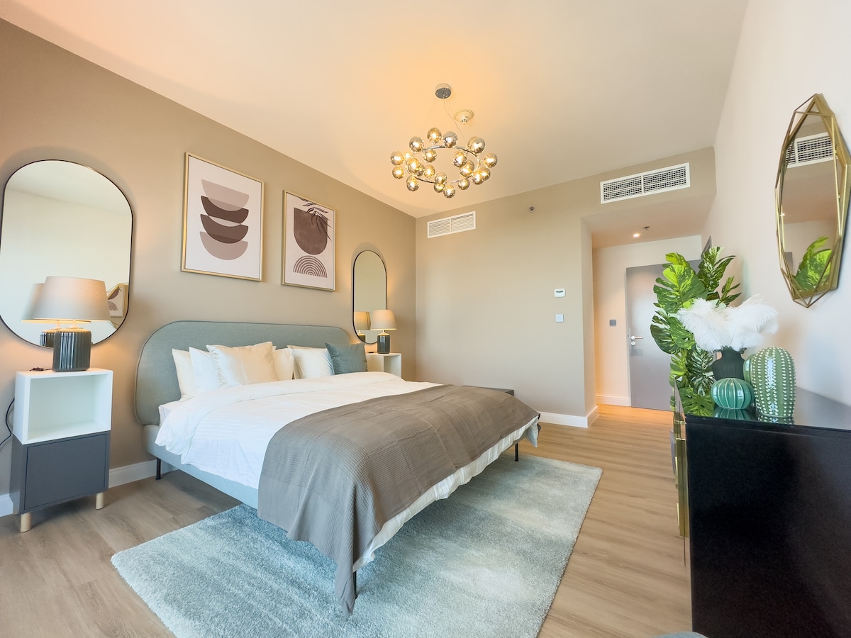 A spacious bedroom is highlighted by a large bed with neutral bedding and an elegant headboard. Decorative mirrors adorn the walls, and a unique light fixture hangs from the ceiling. A bedside table and modern dresser complement the contemporary design, while a green plant adds a touch of nature.