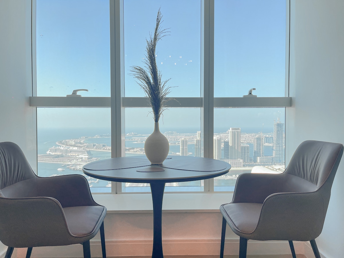 A modern seating area includes two stylish chairs positioned around a round table. A vase with decorative foliage is centered on the table, while expansive windows showcase views of Dubai Harbor and Bluewaters, providing an abundance of natural light.