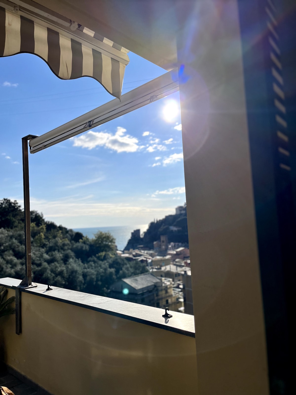 A vibrant view captures the coastline under a clear blue sky, framed by a window with a retractable awning. Sunlight creates a warm glow, illuminating the distant village and lush greenery that contrast with the serene sea.