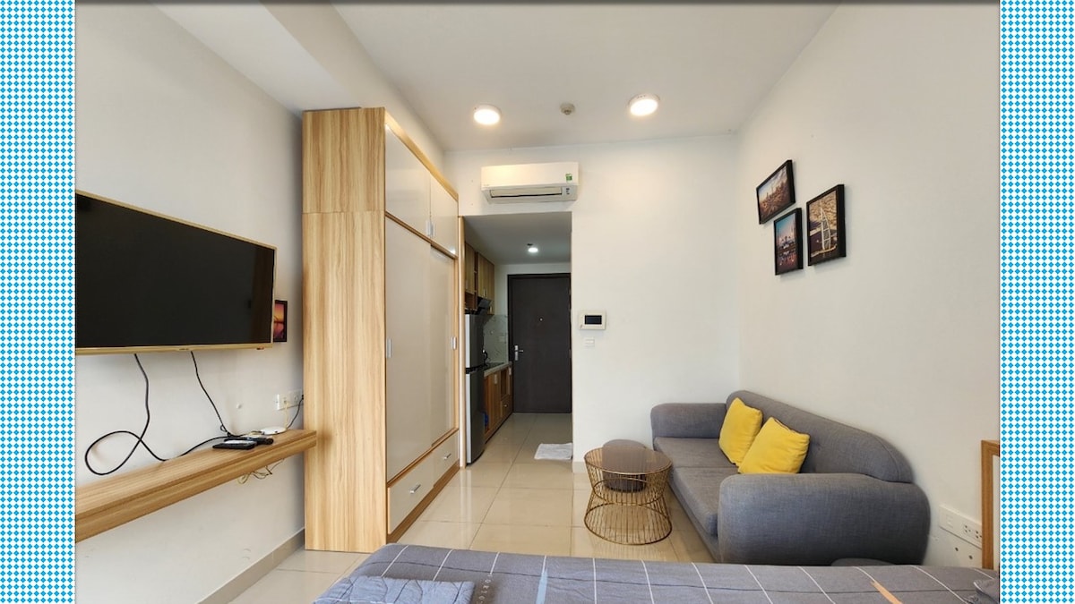 A compact living space features a grey sofa with yellow cushions, positioned beside a round coffee table. A wall-mounted television is visible, along with light-colored cabinetry and a kitchenette area in the background. Natural light fills the room, enhancing the modern design.