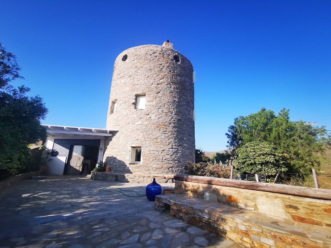 The exterior of the stone windmill is prominently displayed against a clear blue sky. The circular structure features several windows, and the entrance is framed by a small stone wall. Lush greenery surrounds the property, enhancing the serene environment.