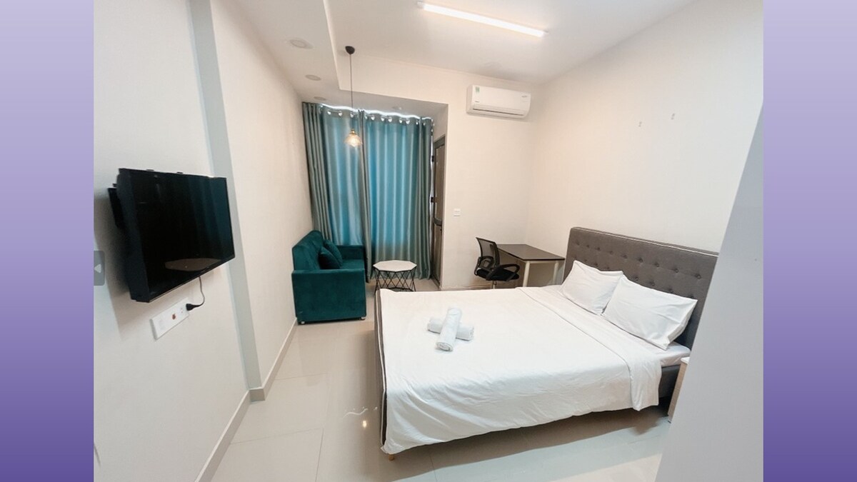 A modern room features a spacious bed with fresh linens, accompanied by a seating area with a green couch. A desk and chair are positioned near a window covered by light curtains. A television is mounted on the wall, enhancing the room's functionality.