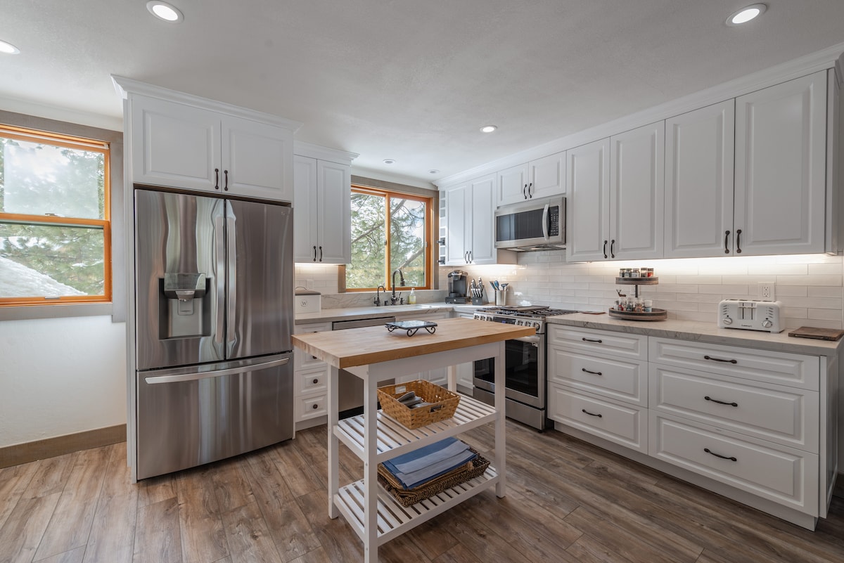 A spacious kitchen features white cabinetry and stainless steel appliances, including a large refrigerator, oven, and microwave. A central island with a wooden top provides additional workspace, while natural light pours in through two windows, illuminating the counters and cooking tools.