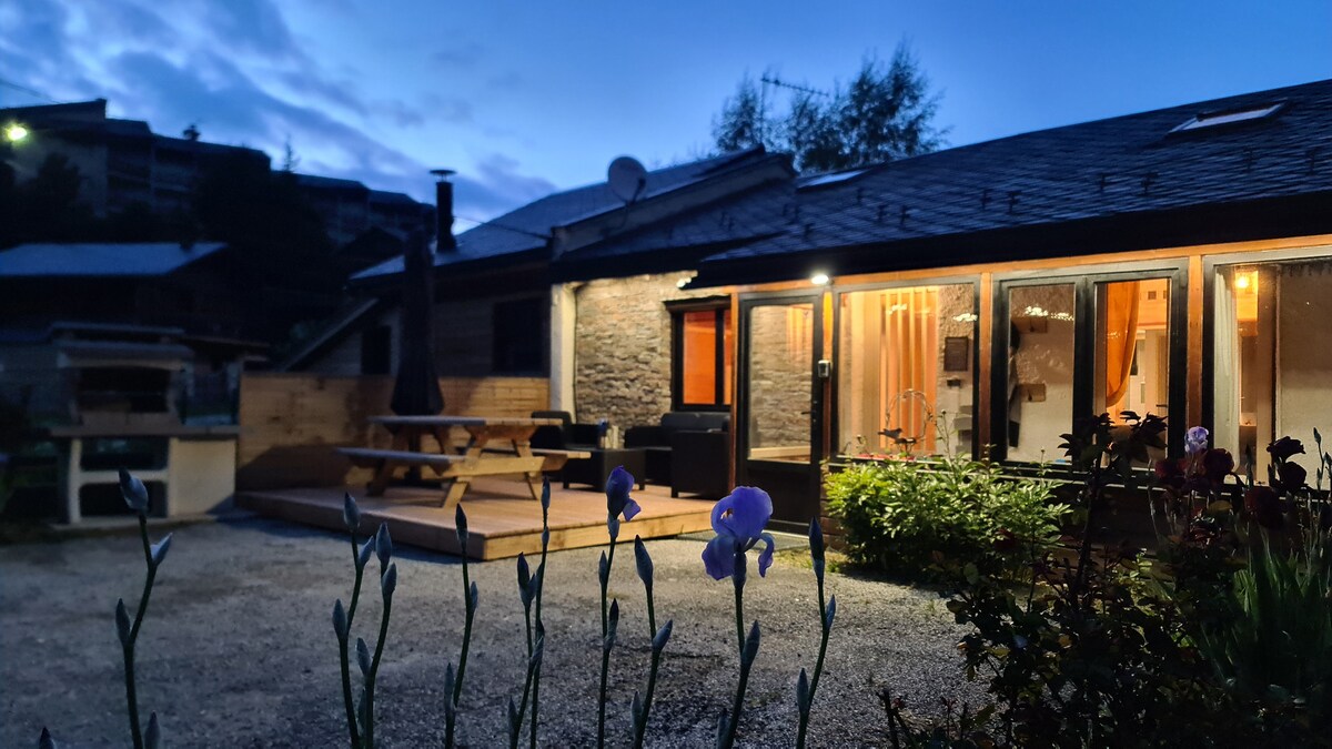 The exterior of the home is depicted during twilight, highlighted by soft lighting. A spacious terrace with a barbecue area and outdoor seating is visible. Flowers, including irises, frame the scene, adding a natural touch to the surrounding garden.
