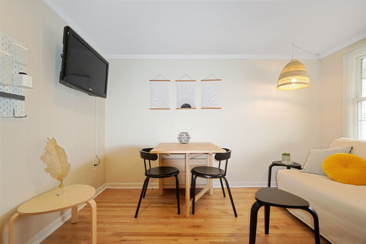 A stylish living area showcases a light wooden table surrounded by four black chairs. The space features a sleeper sofa adorned with decorative pillows, a mounted television, and two framed art pieces on the beige wall. A pendant light and small side table enhance the cozy atmosphere.