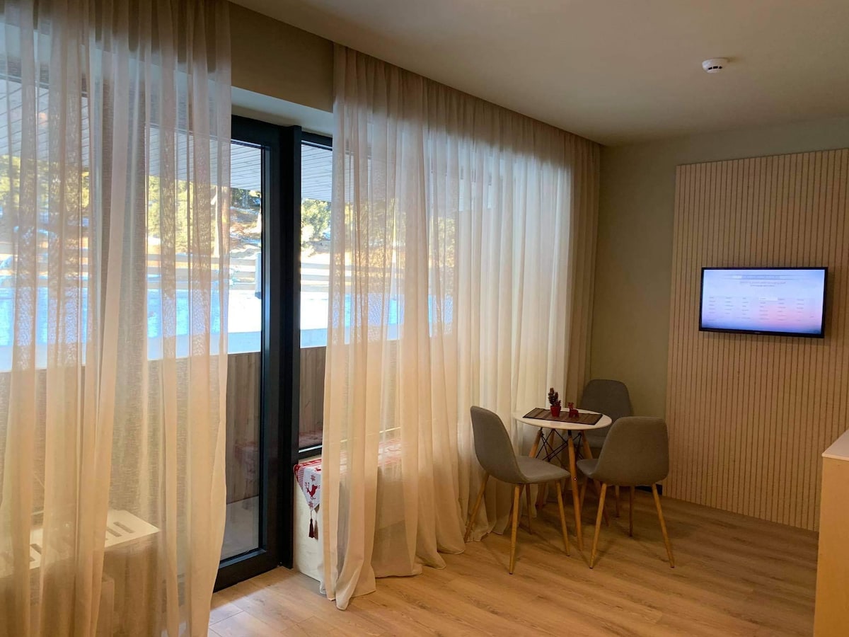 A cozy corner features a small table with two light grey chairs, set against large glass doors draped in sheer curtains. Natural light floods the space, highlighting the wooden flooring. A wall-mounted television is positioned nearby, complementing the modern design.