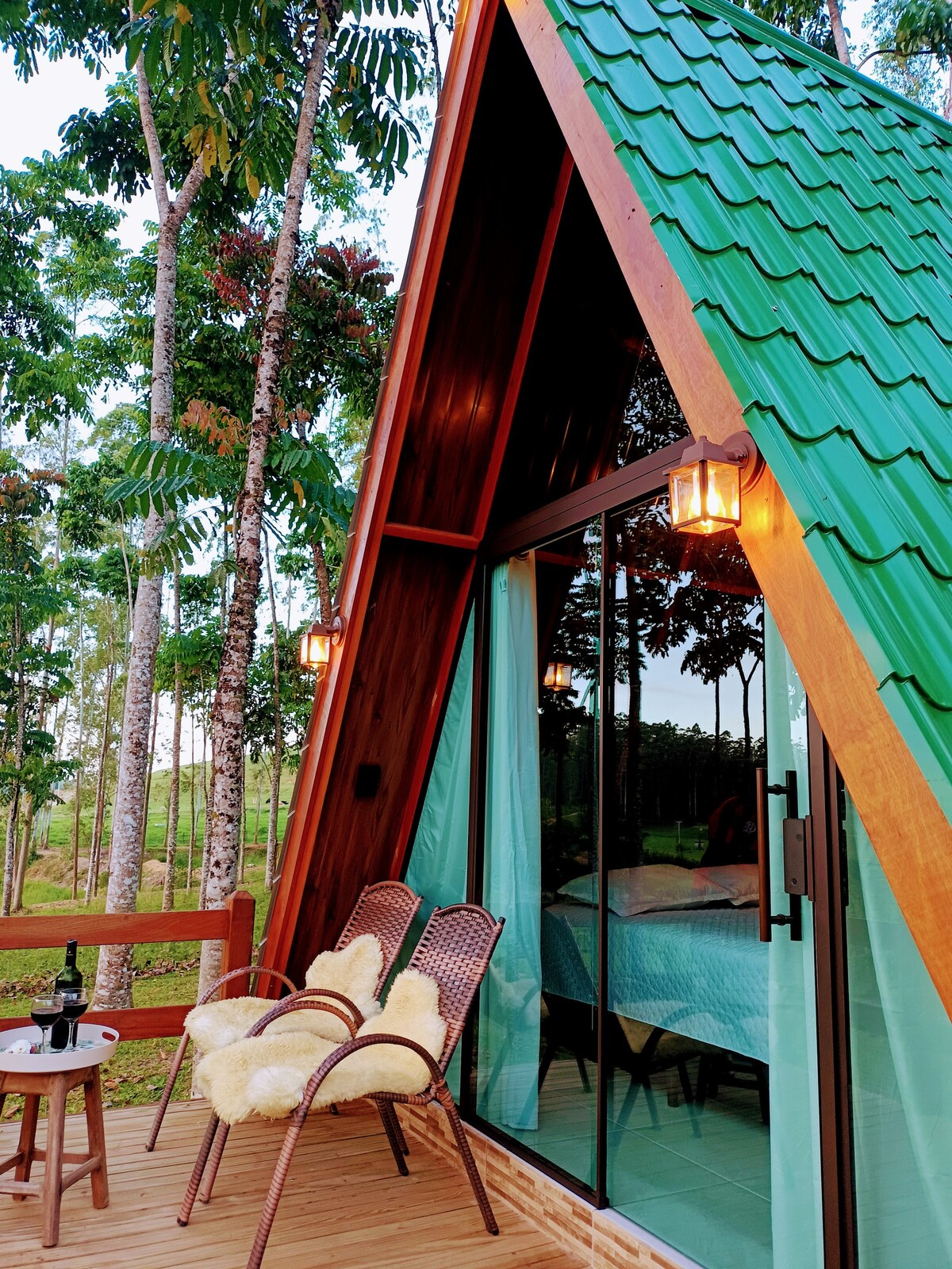 A charming chalet exterior features an A-frame design with a green roof, framed by lush trees. A private deck includes two woven chairs adorned with soft throws, inviting relaxation. Glass doors reveal the cozy interior, enhancing the connection with the surrounding natural beauty.
