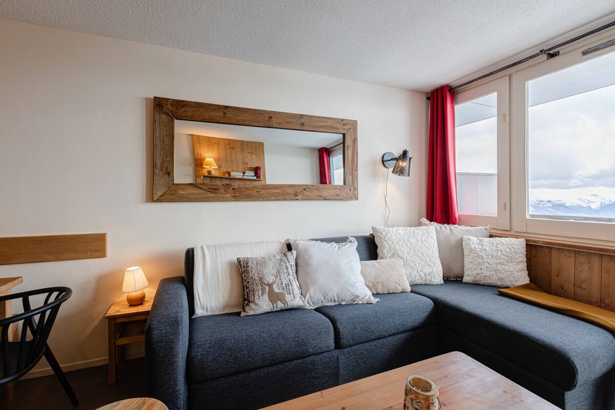 A spacious living area is highlighted by a comfortable gray sectional sofa adorned with decorative pillows. A wooden mirror reflects soft lighting, complemented by a small lamp on a side table. Large windows offer views of the surrounding mountains, with red curtains framing the scene.