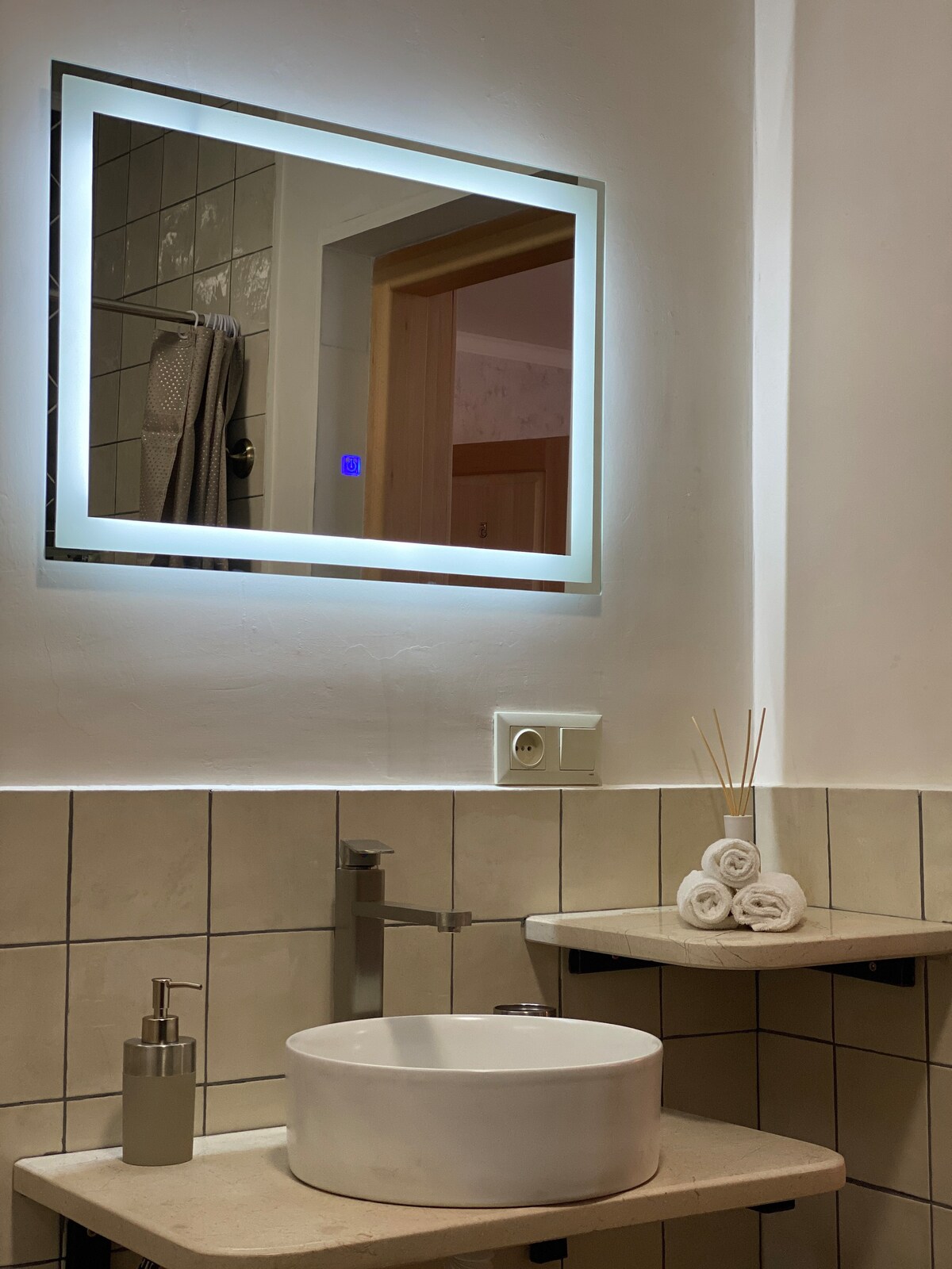 A well-lit bathroom features a modern sink with a circular basin positioned on a minimalist countertop. Above, a backlit mirror enhances visibility. Neatly arranged rolled towels and a decorative diffuser add to the space's functionality and understated elegance.