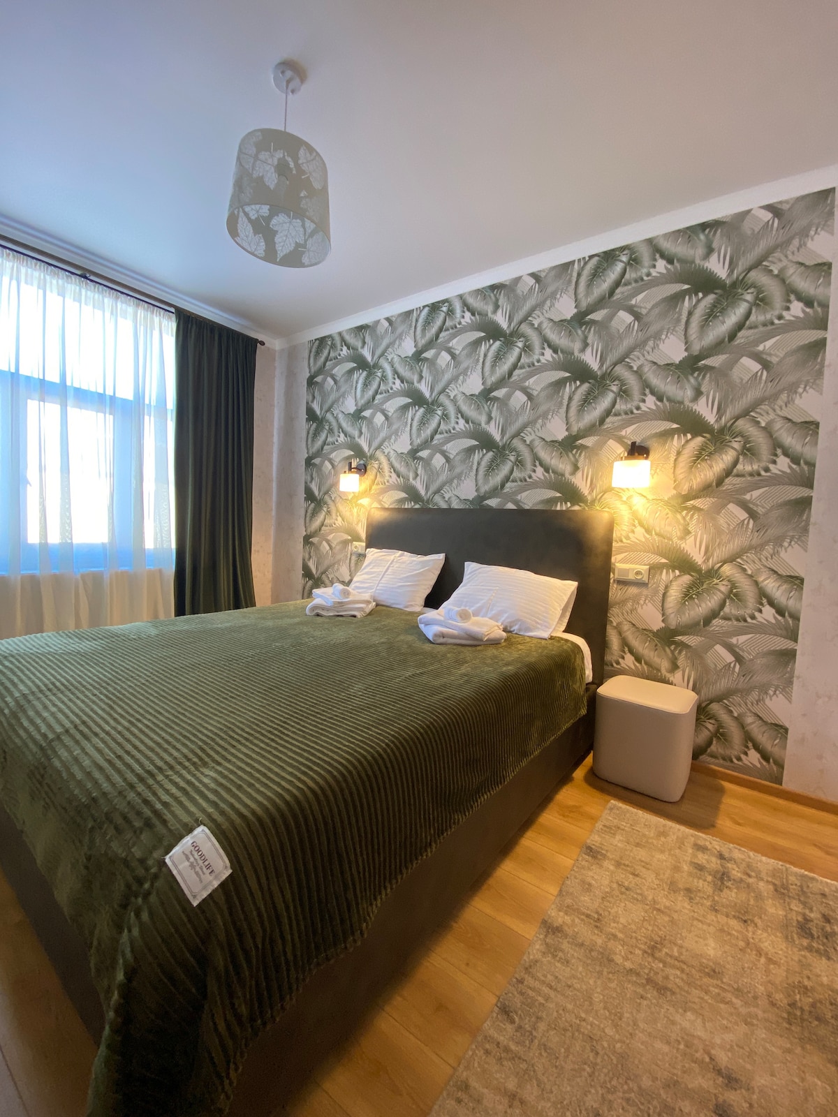 A sleeping area features a spacious bed dressed in an olive green coverlet, flanked by two sconces providing soft lighting. The walls are adorned with a leafy wallpaper pattern, and a window draped in dark curtains allows natural light to filter through.