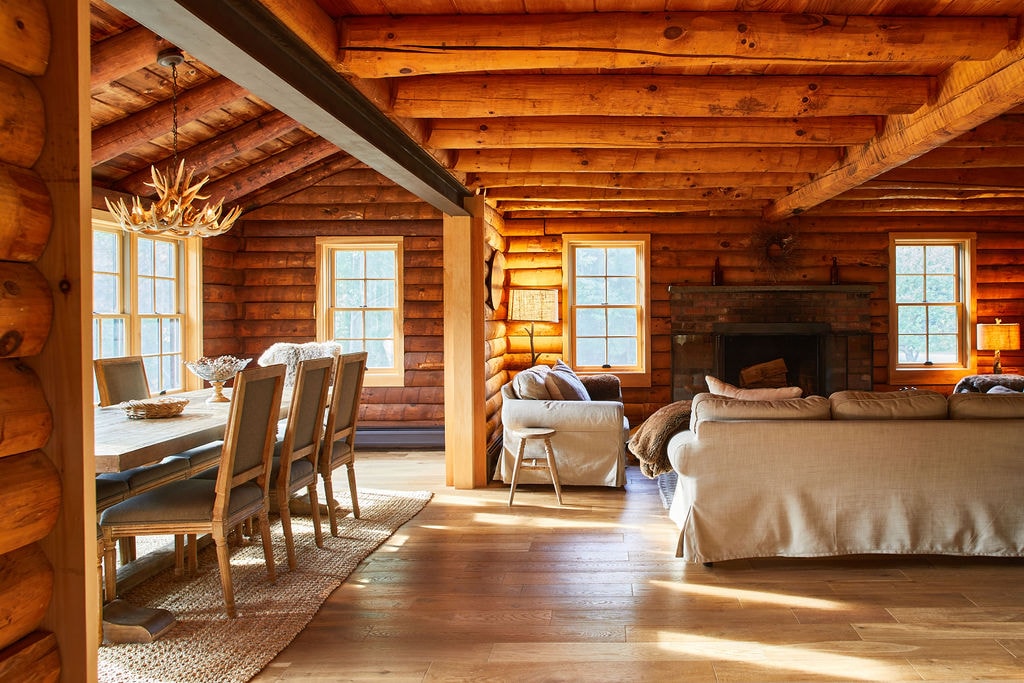 The living area features warm wooden walls and ceilings, enhancing the rustic charm. A spacious dining table with light-colored chairs is positioned near large windows, while a cozy seating arrangement invites relaxation. Natural light streams in, highlighting the texture of the wood and furnishings.