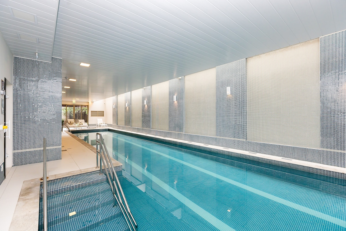 An indoor heated pool is surrounded by a sleek tiled environment, featuring a walk-in staircase for easy access. Soft lighting enhances the serene atmosphere, while lounge chairs are provided for relaxation along one side of the pool.