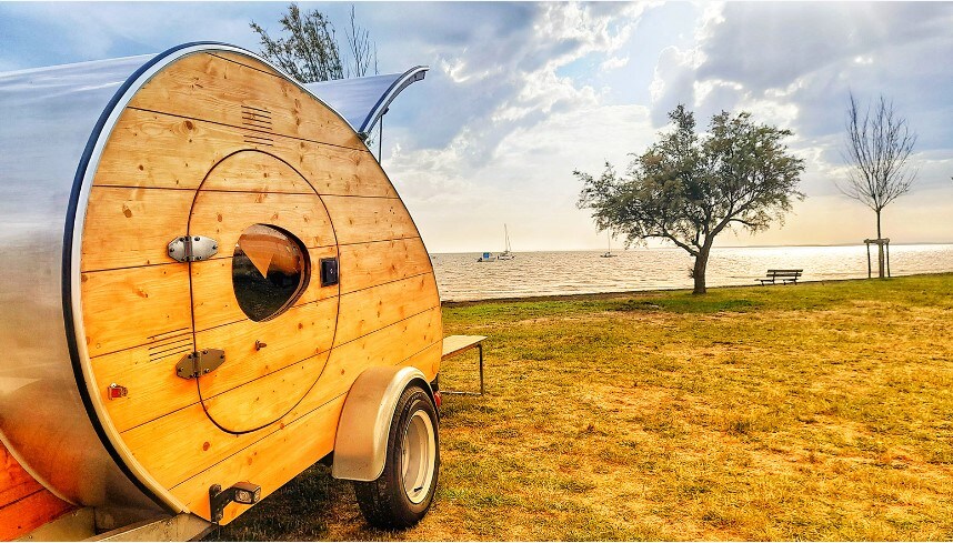 A teardrop caravan with a wooden exterior is positioned on a grassy area near the water's edge. The scene features a calm lake in the background, with sailboats visible on the horizon. A tree and a bench complete the serene outdoor setting.