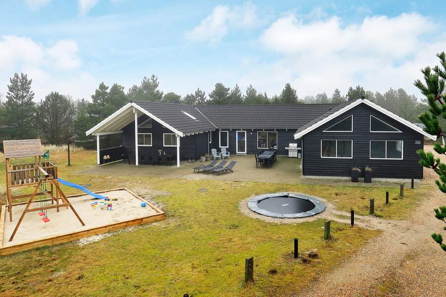 A spacious summer house is set amidst a natural landscape, featuring a large outdoor area with a sandpit and a play structure. A trampoline is positioned nearby, while several seating options are arranged on the patio. Trees line the background, providing a sense of privacy.