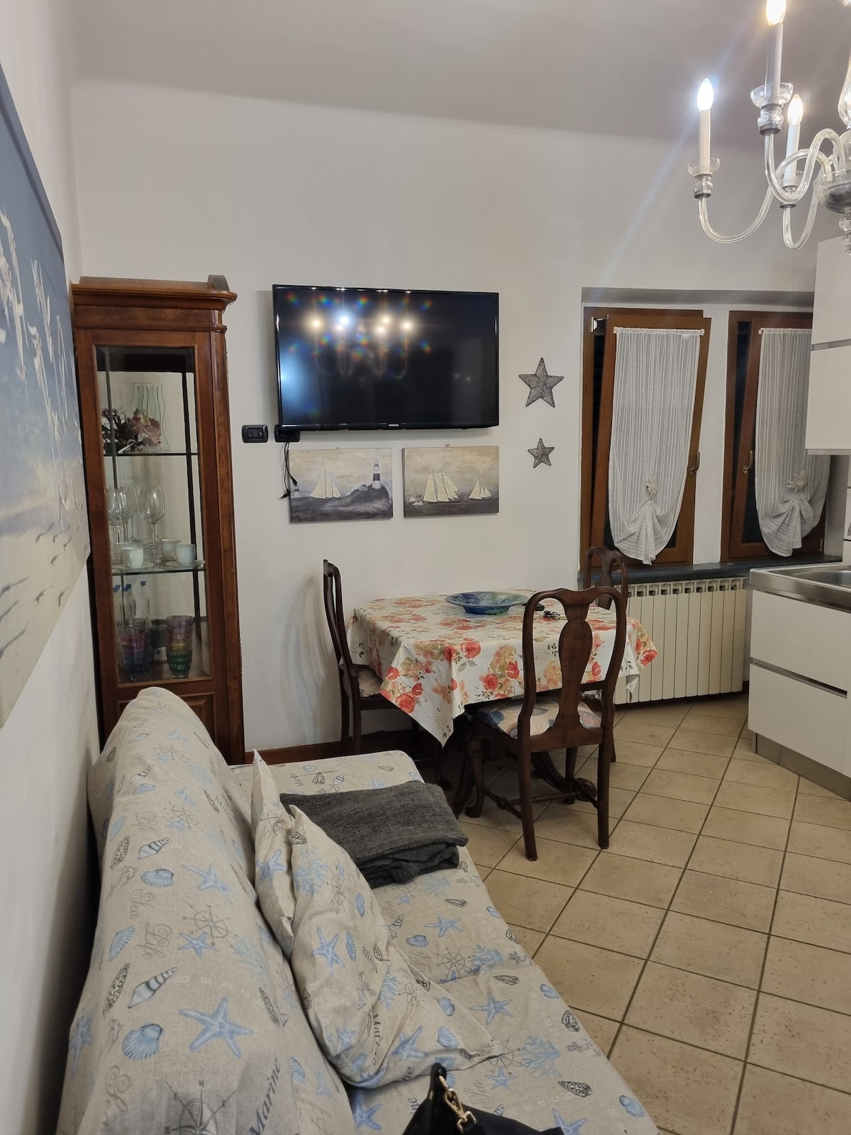 A cozy living area is presented, featuring a light-colored sofa adorned with nautical-themed patterns. A wooden dining table is set with floral placemats, accompanied by matching chairs. A television mounted on the wall adds to the entertainment options, with a glass cabinet displaying decorative items.