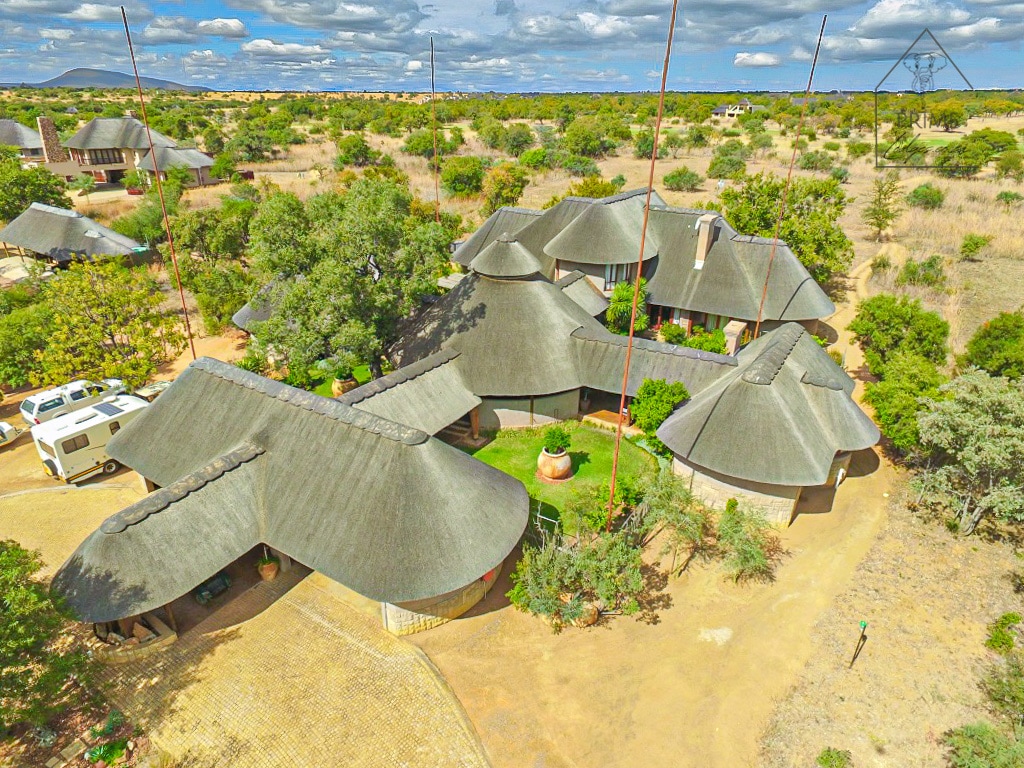 An aerial view of Cherry Creek lodge showcases a unique layout of thatched-roof buildings set in a natural landscape. Lush greenery surrounds the central courtyard, while pathways connect the structures, emphasizing the spaciousness of the property against the backdrop of the African bushveld.
