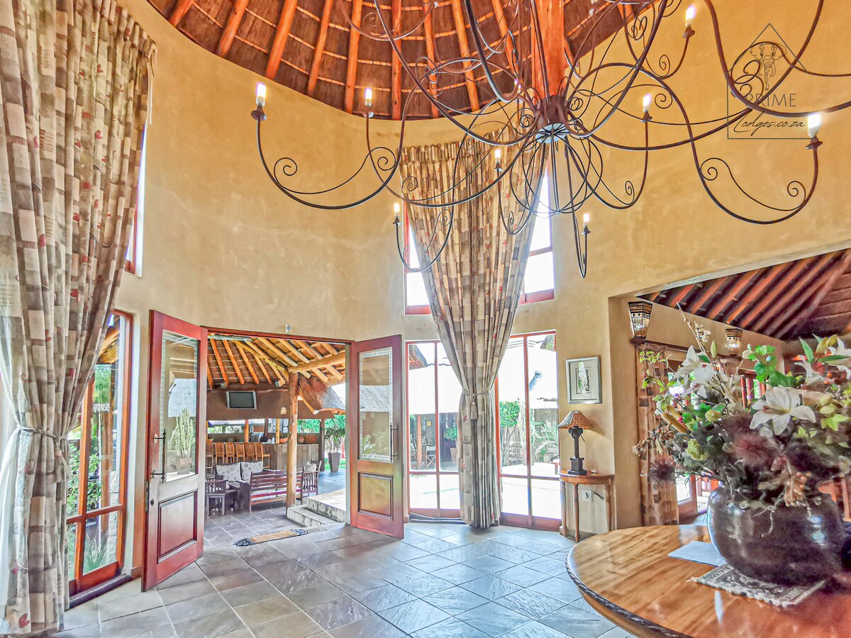 A spacious entryway features high, rounded ceilings and large windows that invite natural light. Elegant curtains frame the windows, while a decorative chandelier adds character. A round table with subtle decor sits in the foreground, providing a welcoming transition to the outdoor gathering area visible beyond.