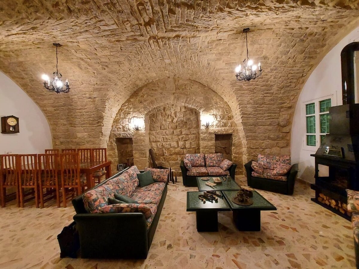 A spacious sitting area showcases traditional stone walls with arched ceilings. Two patterned sofas and a coffee table are positioned in the center, complemented by wooden chairs in the background. A cozy wood stove stands against one wall, while soft lighting is provided by chandeliers.