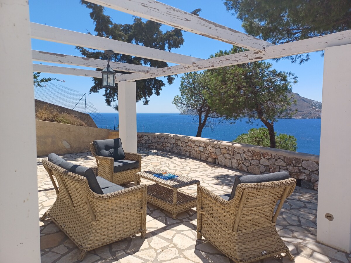 A spacious outdoor seating area features comfortable wicker chairs arranged around a low table. The stone-paved terrace is partially shaded by wooden beams, offering views of the sea and surrounding greenery. Natural light highlights the serene atmosphere, ideal for relaxation.