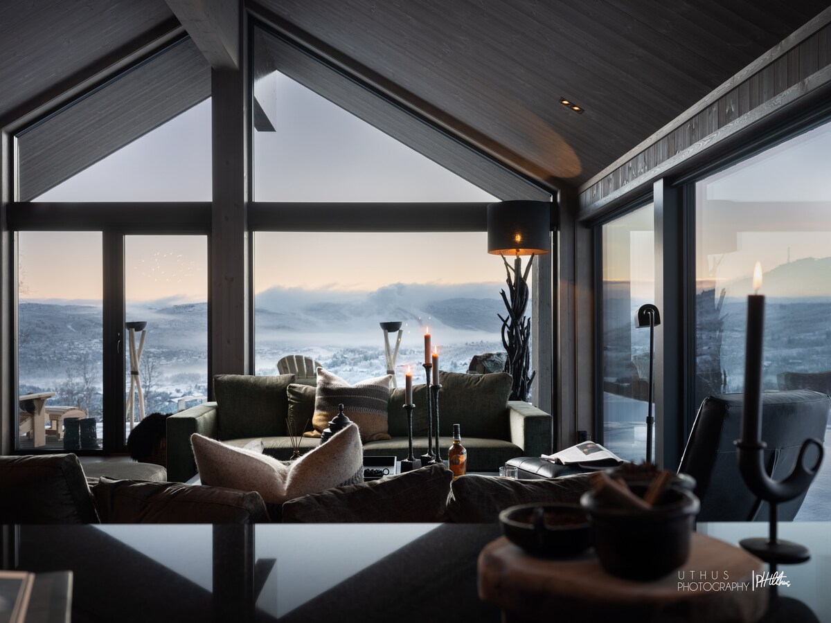 A stylish living area is presented with large panoramic windows, showcasing views of the surrounding landscape. Comfortable seating is arranged around a central coffee table, with candles providing soft illumination. The modern design is enhanced by natural elements and a cozy atmosphere.