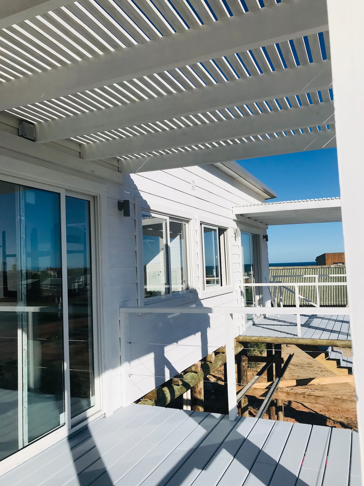 The image displays a bright exterior view of the cabin's deck, featuring white wooden siding and multiple large windows. A slatted pergola provides partial shade, and the deck is constructed with light grey flooring, enhancing the space's openness and connection to the surroundings.