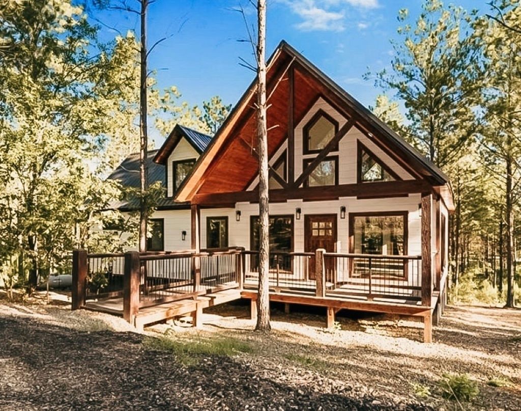 A modern cottage is framed by tall trees, featuring a sloping roof and large windows. A spacious wooden deck extends from the front entrance, offering ample space for outdoor relaxation. Natural surroundings enhance the secluded setting, promoting a serene atmosphere.