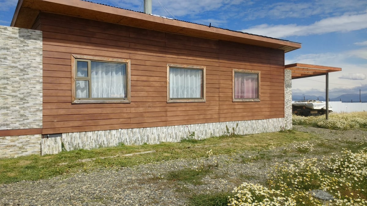 A single-story house features wooden paneling and a stone accent base. Three windows with light-colored curtains are visible, allowing for natural light. A covered porch extends from the structure, and the surrounding area is filled with greenery and wildflowers, creating a serene outdoor environment.