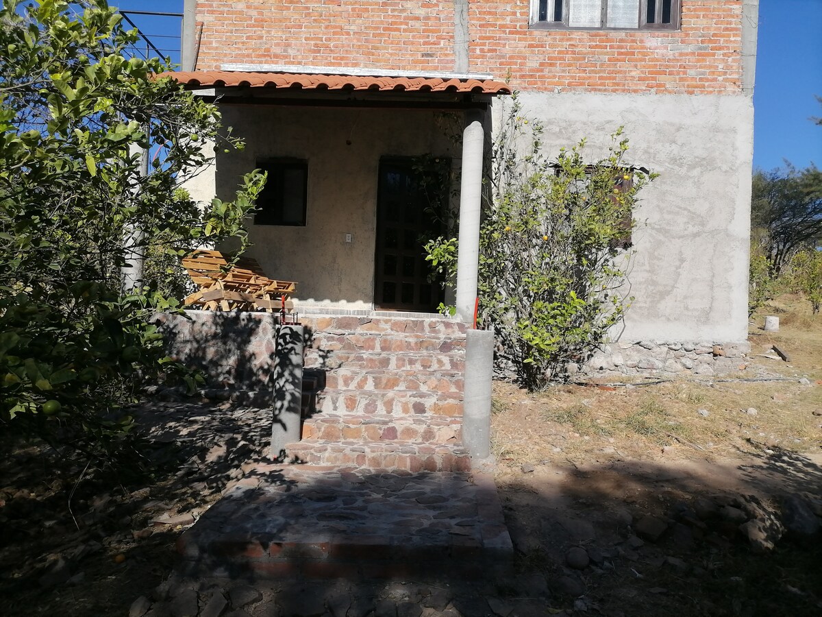The entrance of a rustic home is framed by a stone pathway leading to a covered porch. Surrounding greenery includes small shrubs and trees, providing a natural setting. The house features a mix of stone and brick materials, contributing to its earthy ambiance.