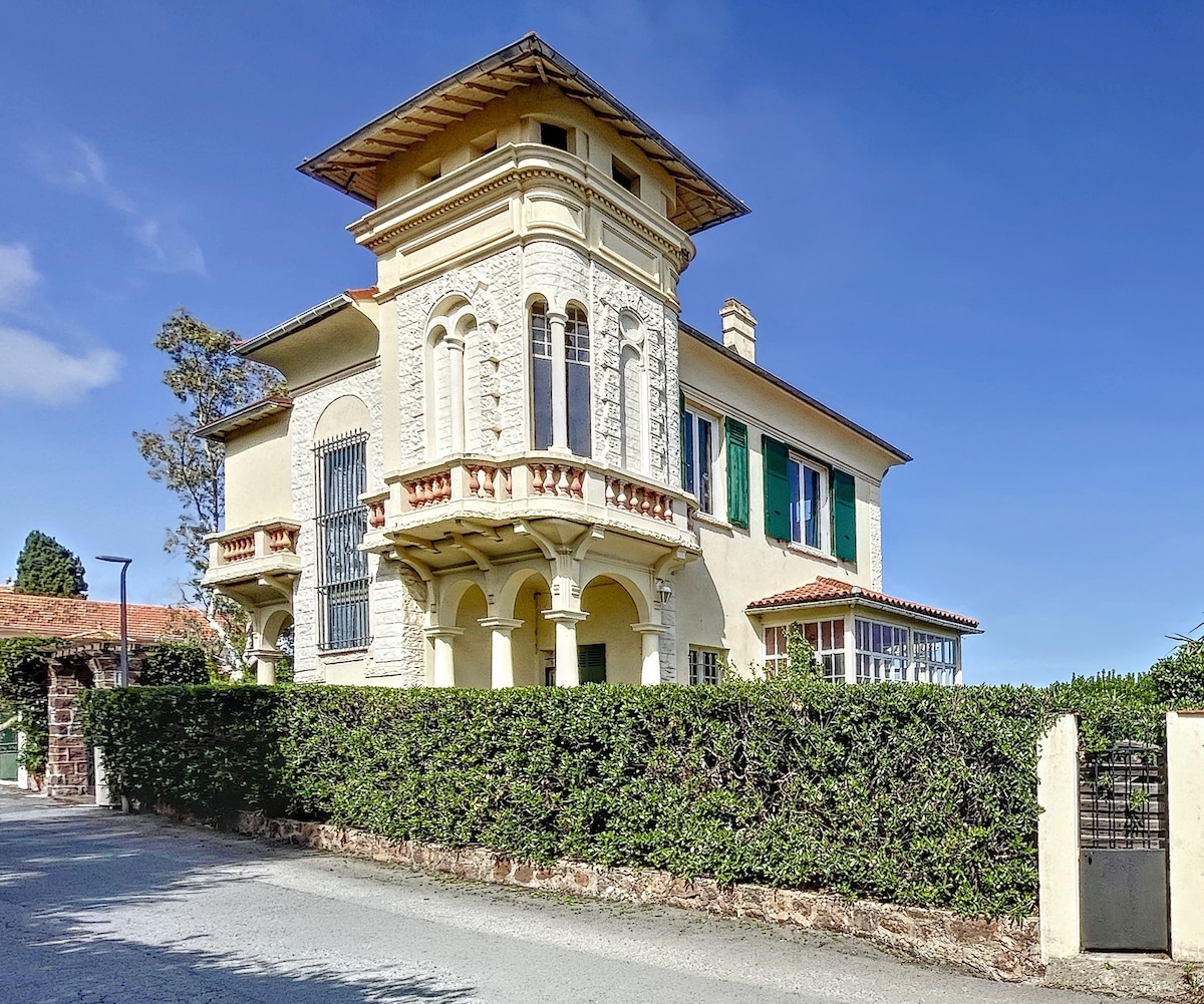 The villa, showcasing a classic architectural style, features intricate stonework and a prominent balcony. Green shutters adorn the windows, while a lush hedge surrounds the property. The building is set against a clear blue sky, highlighting its grandeur and inviting presence.