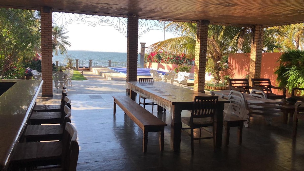 An open-air terrace offers seating with a large wooden table and various chairs. The space features views of the sea and is framed by tropical plants and vibrant flowers. A swimming pool and lounge area are visible in the background, inviting outdoor relaxation.