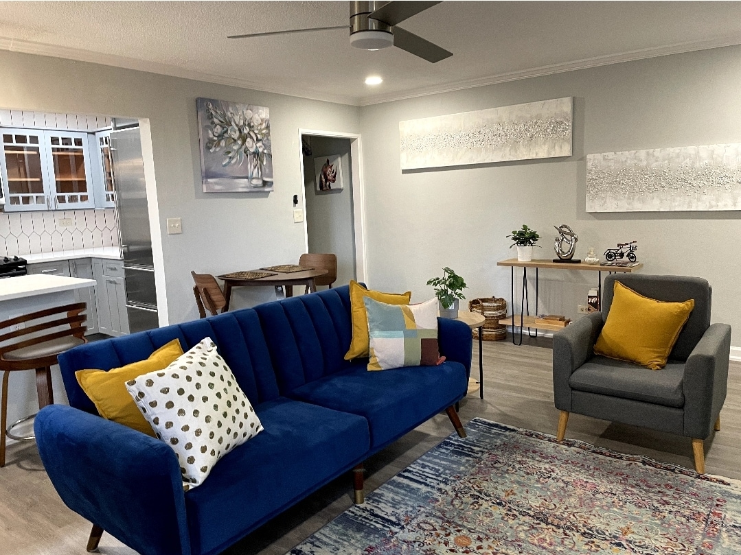 A cozy living area is furnished with a blue sofa and a gray armchair, accented by colorful pillows. Artwork is displayed on the wall, and a small plant adds a touch of greenery. A dining table is visible in the background, leading to a well-lit kitchen.
