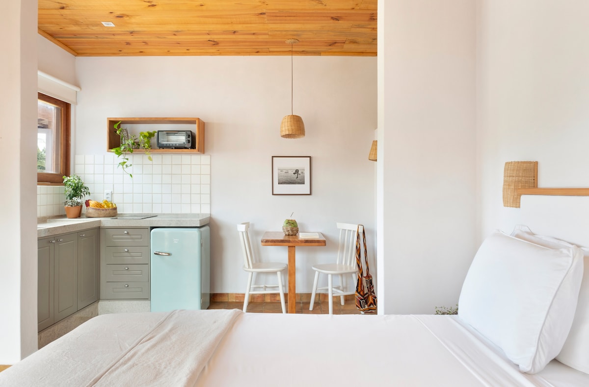 A well-appointed living space features a cozy sleeping area and a kitchenette. The kitchen includes modern appliances and lush greenery, while a wooden dining table with two chairs invites guests to enjoy meals in a bright, airy atmosphere.