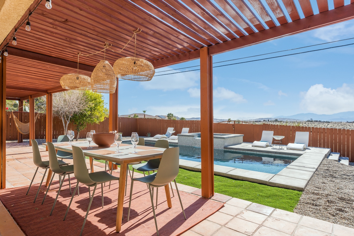 An inviting outdoor dining area features a large table surrounded by chairs, with woven pendant lights above. A saltwater pool is visible nearby, along with lounge chairs positioned on the pool deck. The serene desert landscape and mountains provide a beautiful backdrop.