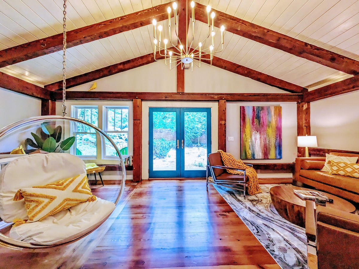A spacious living area features exposed wooden beams and a contemporary chandelier. A suspended bubble chair adds a unique touch, while large blue doors open to the outdoor views. A cozy sectional sofa and a colorful art piece create an inviting atmosphere.