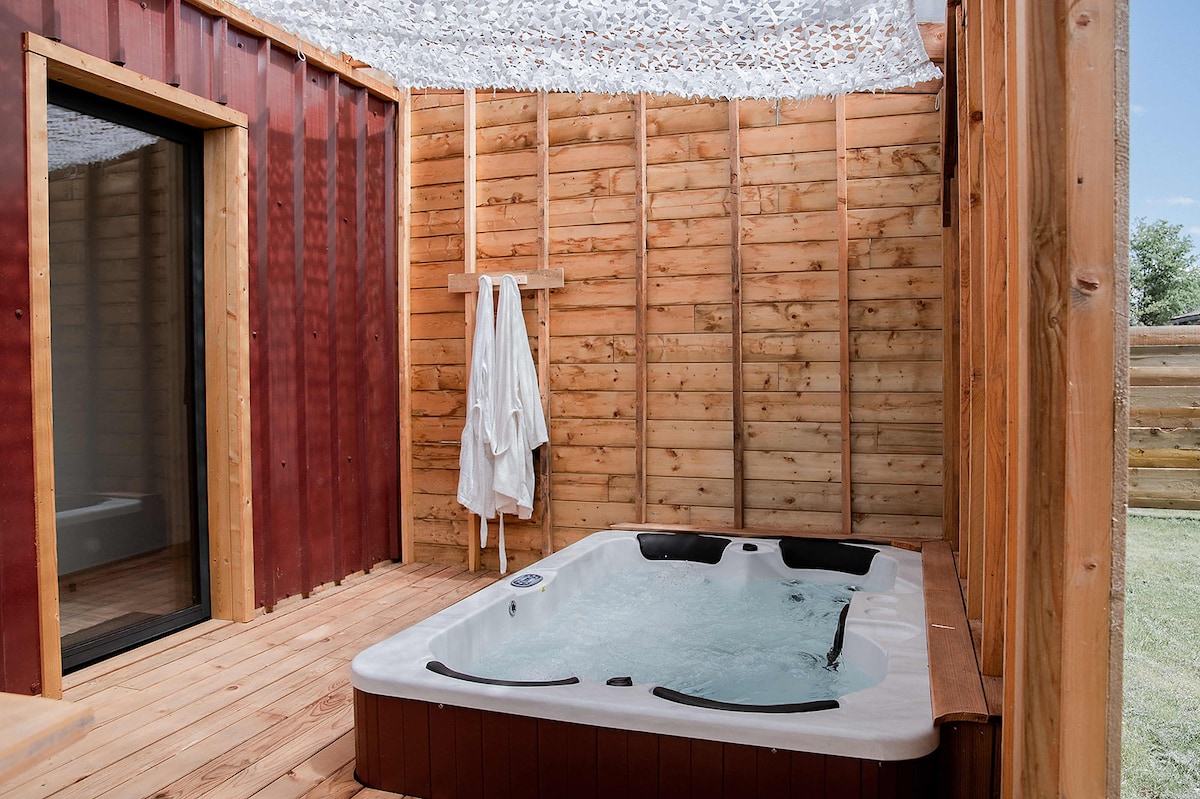 A private spa area is enclosed by wooden walls, featuring a hot tub designed for four people. Light streams in through a glass door, and two white bathrobes are hung nearby, creating a serene and inviting atmosphere for relaxation.