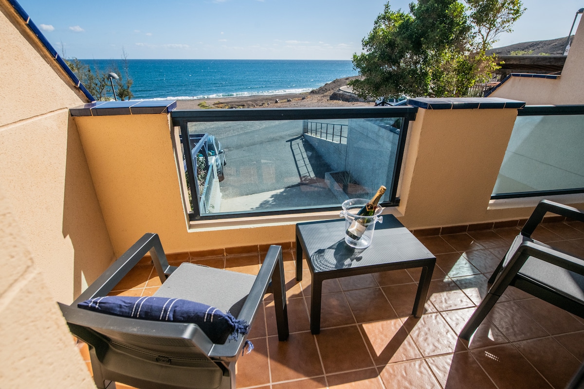 A private balcony offers a clear view of the ocean, with sunlight reflecting off the water. Two chairs are positioned around a small table, which holds an ice bucket. The coastal scenery is easily accessible through the glass railing.
