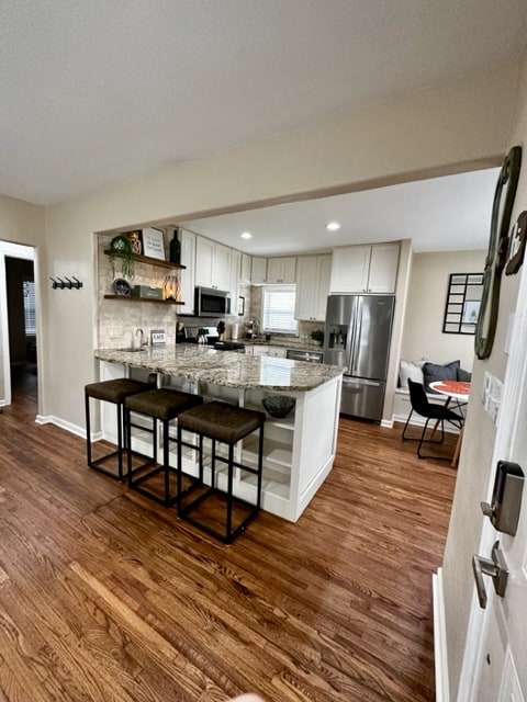 The kitchen area features a spacious island with three bar stools and a polished countertop. Modern appliances, including a refrigerator and microwave, are present, and a dining table is visible in the background. Natural light enters through the window, enhancing the inviting ambiance.
