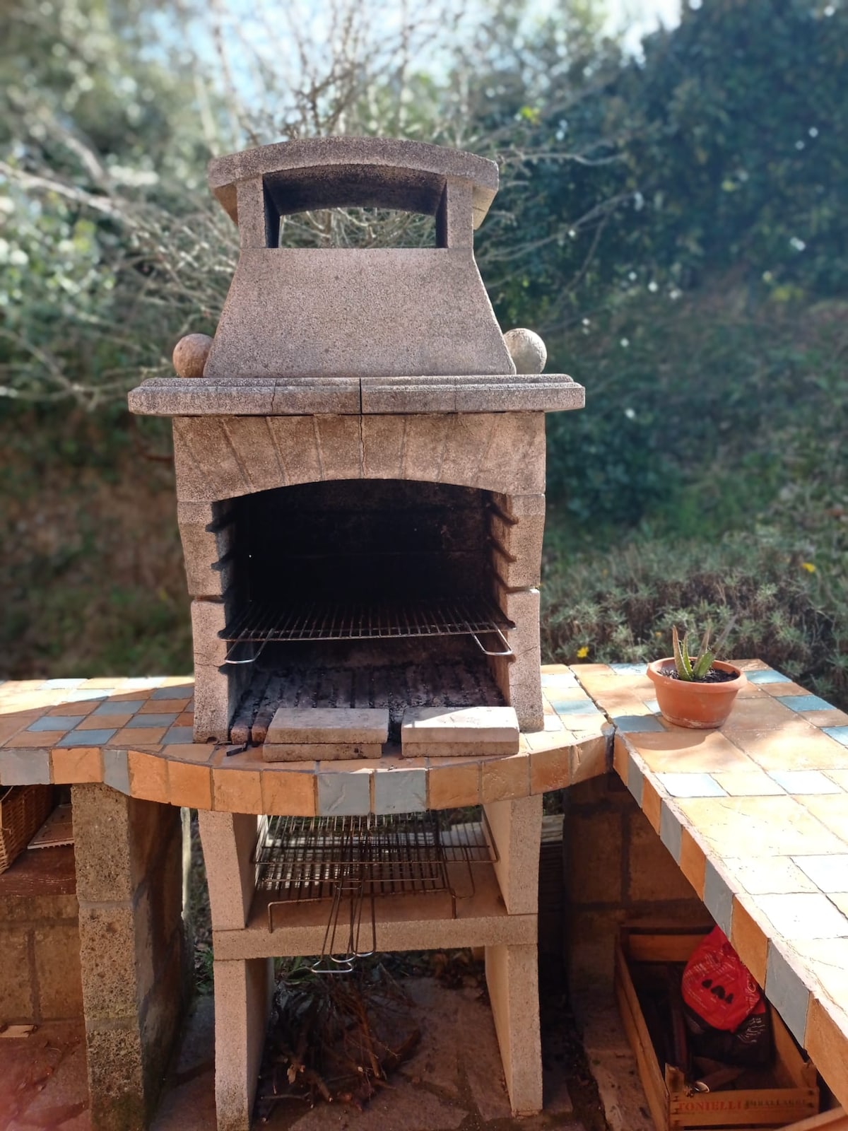 A stone barbecue grill is positioned on a tiled outdoor countertop, featuring a traditional chimney and grilling area. A small potted plant is visible nearby, surrounded by greenery in the background, enhancing the outdoor setting.