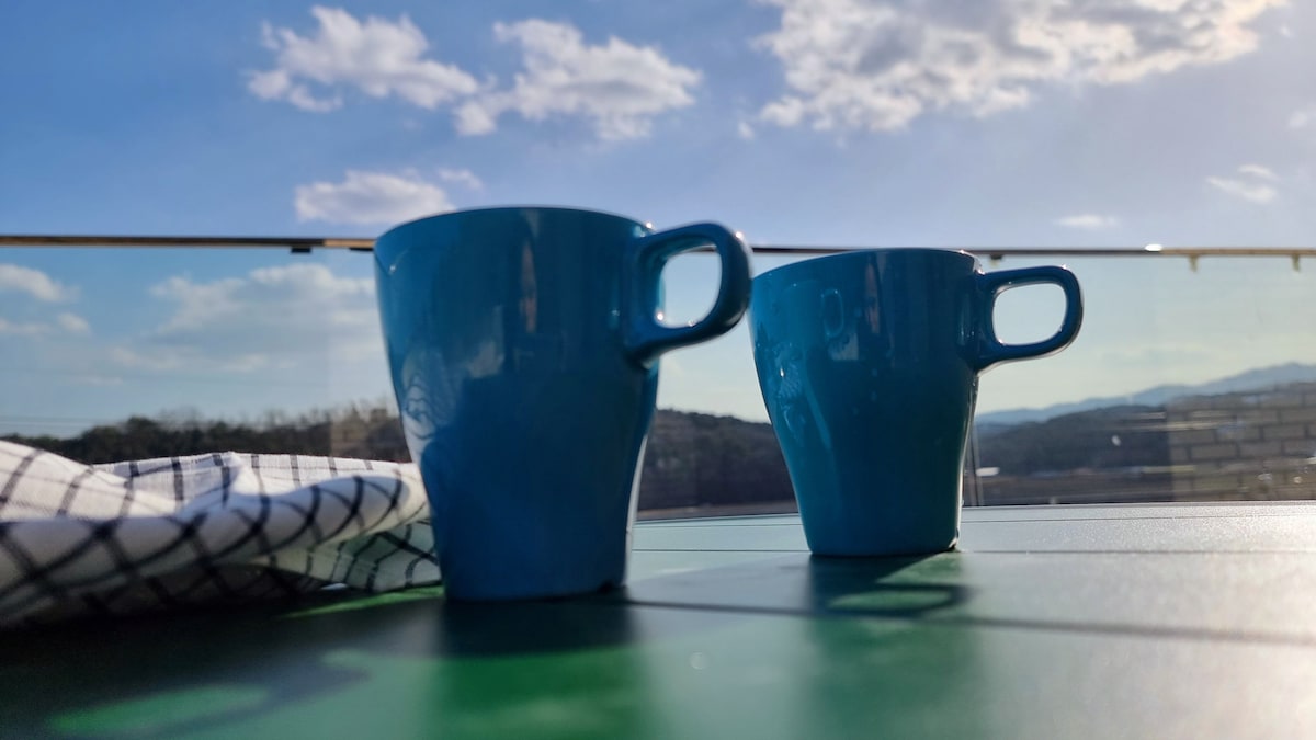 Two blue ceramic mugs sit on a green table, with a checkered cloth nearby. The background features a clear sky and distant mountains, creating a serene outdoor scene.