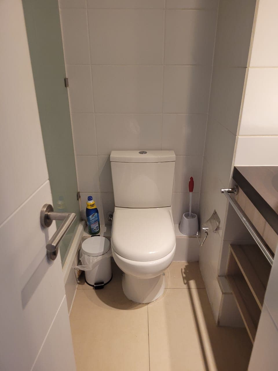 A compact bathroom features clean white tiles with a modern toilet positioned next to a small wastebasket and a cleaning supply container. A toilet brush holder is also visible, and the sink area includes a simple countertop with shelves underneath.