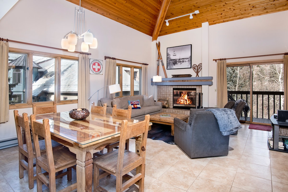 A spacious living area is highlighted by a vaulted wood ceiling and large windows that allow natural light to fill the room. A cozy wood burning fireplace is surrounded by comfortable seating, and a wooden dining table accommodates multiple guests.
