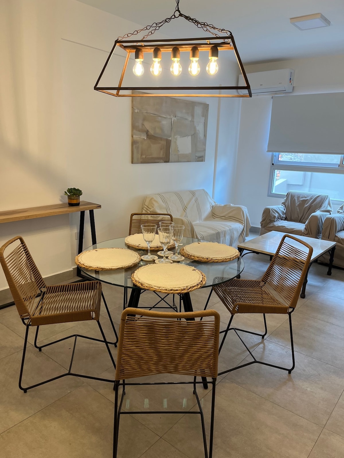 A dining area is presented with a round glass table surrounded by six woven chairs. A modern light fixture hangs above, illuminating the space. Two comfortable armchairs sit nearby, creating a cozy setting. Natural light enters through the window, highlighting the neutral decor.