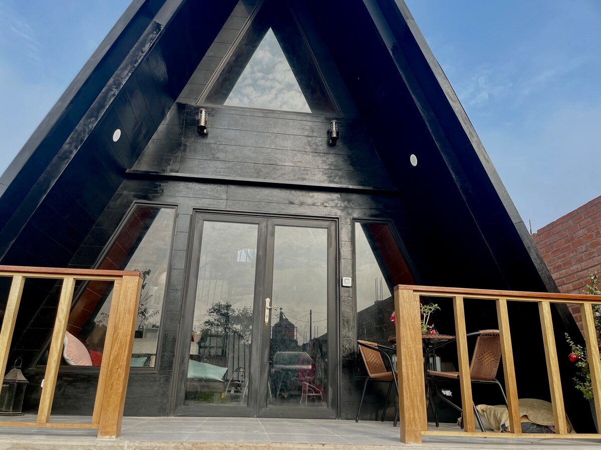 The exterior of the cabin features a striking triangular design with a black facade. Large glass doors are set beneath a sloped roof, flanked by two wall-mounted lights. A wooden railing surrounds a small porch with outdoor seating, creating a welcoming entryway.