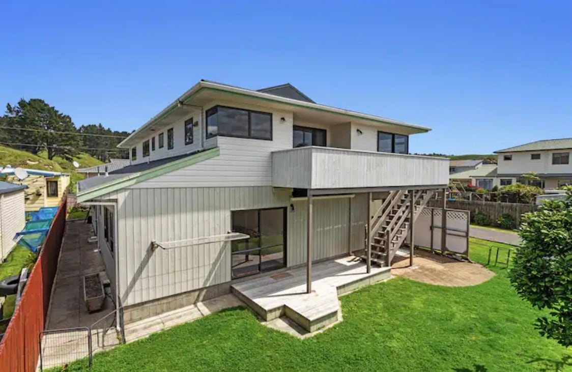 A large two-story beach house is depicted, featuring an elevated deck with stairs leading down to a grassy yard. Large windows provide natural light, and the surrounding area includes other residential buildings. The structure is finished in light-colored materials, creating a spacious appearance.