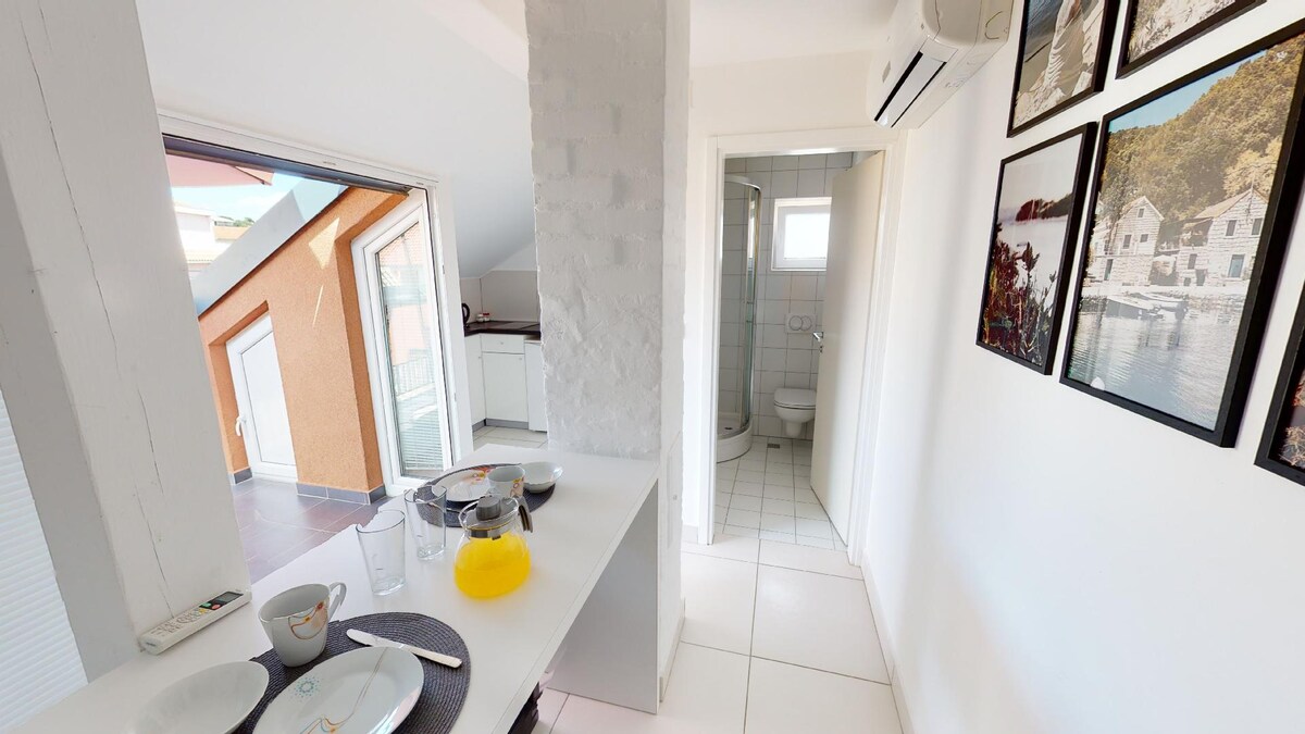 A bright corner of the apartment features a dining table set for breakfast, with plates and glasses arranged on a white surface. The adjoining space shows an open kitchen area and a bathroom door visible in the background, while natural light enters through a nearby window.