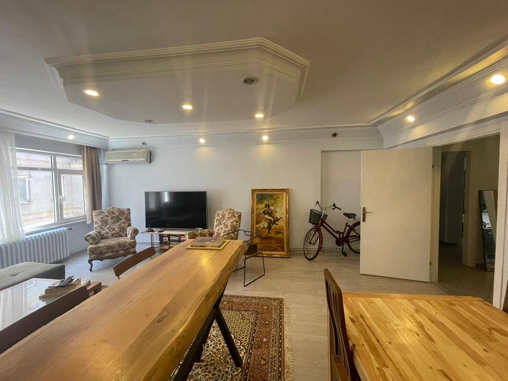 A spacious living area is presented with a large wooden dining table and comfortable seating arrangements. A vibrant area rug adorns the floor, while a television is positioned on one wall. Natural light enters through large windows, and a bicycle is parked nearby.