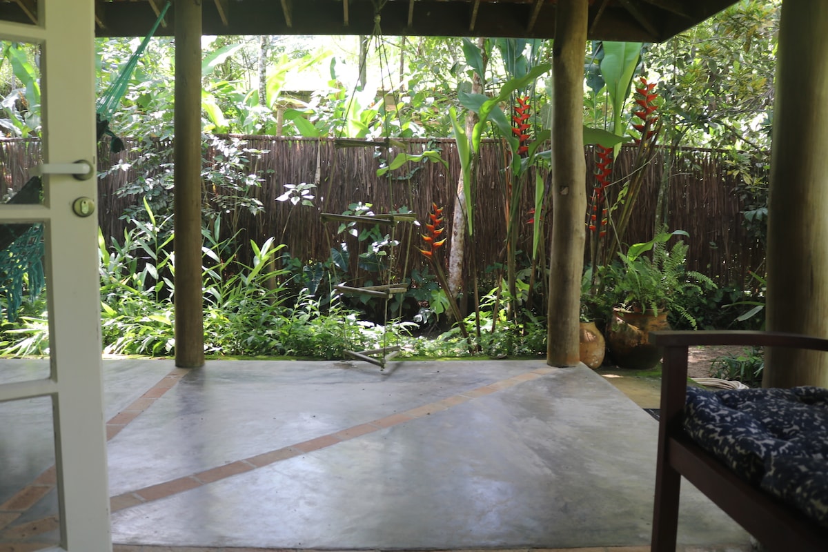 A covered outdoor area is presented, featuring a concrete floor and wooden posts. Lush greenery surrounds the space, with tall plants and vibrant flowers visible in the background. A simple wooden chair sits to the side, enhancing the natural ambiance.