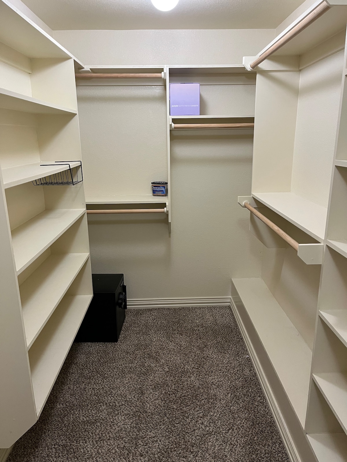 A spacious closet featuring multiple open shelves and hanging rods. The light-coloured walls enhance the sense of space, while a carpeted floor offers comfort. A small black storage box is visible on the ground, and a single light bulb illuminates the area.