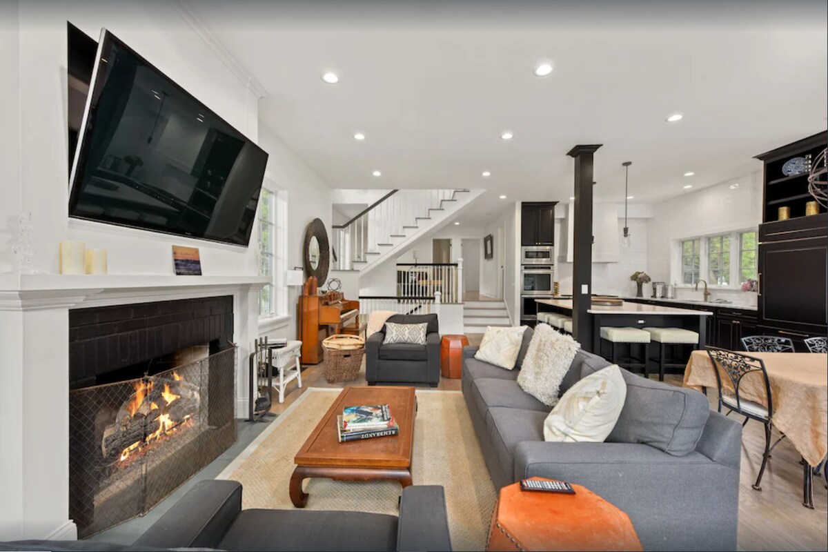 An open concept living area features a cozy wood-burning fireplace, a large television mounted above the mantel, and comfortable seating arrangements. The space transitions into the dining area and kitchen, showcasing modern appliances and natural light from windows.